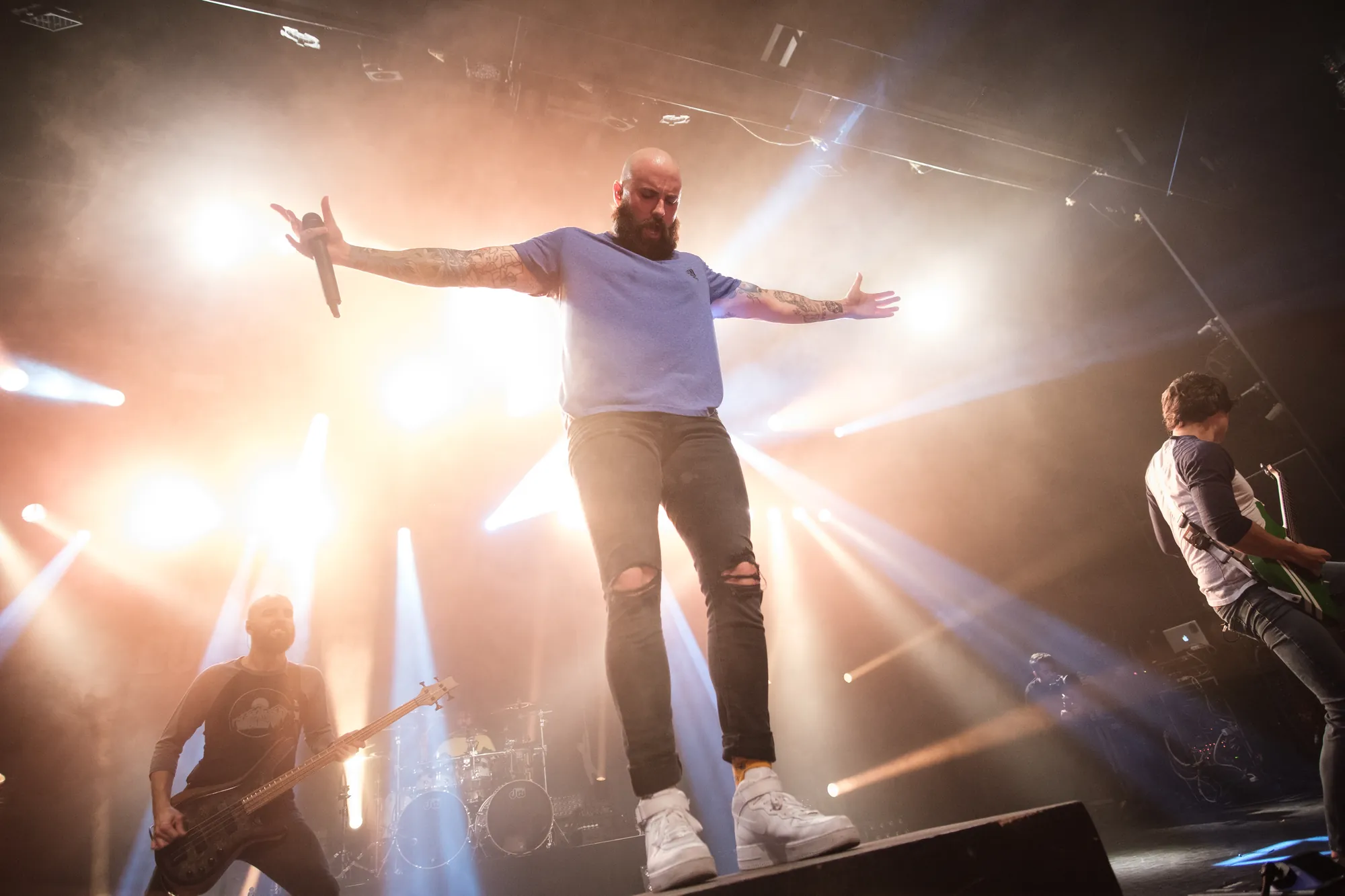 Tattooed vocalist with arms spread wide on stage, band performing behind him in dramatic warm light