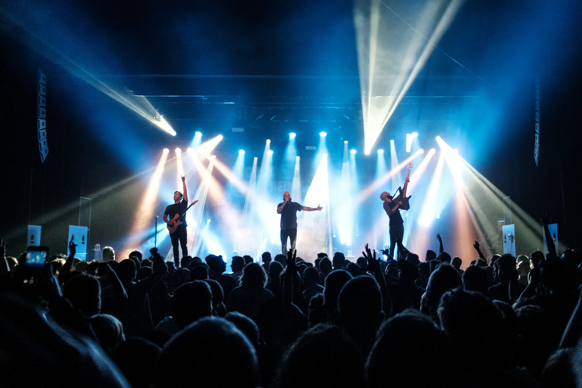 Band performing on stage with blue spotlights, crowd with raised hands in the foreground