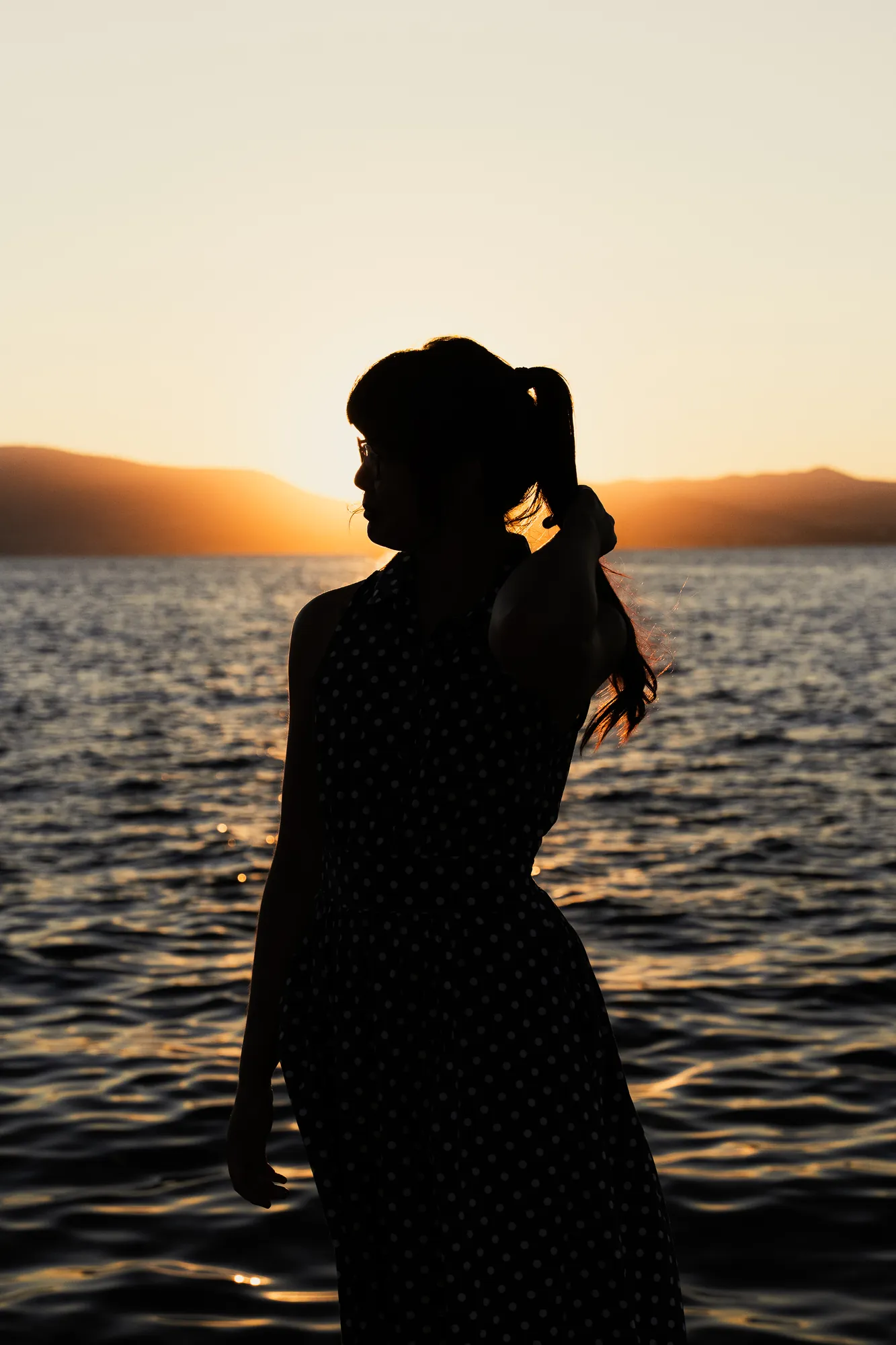 Silhouette of a woman holding her hair against a golden sunset over calm water