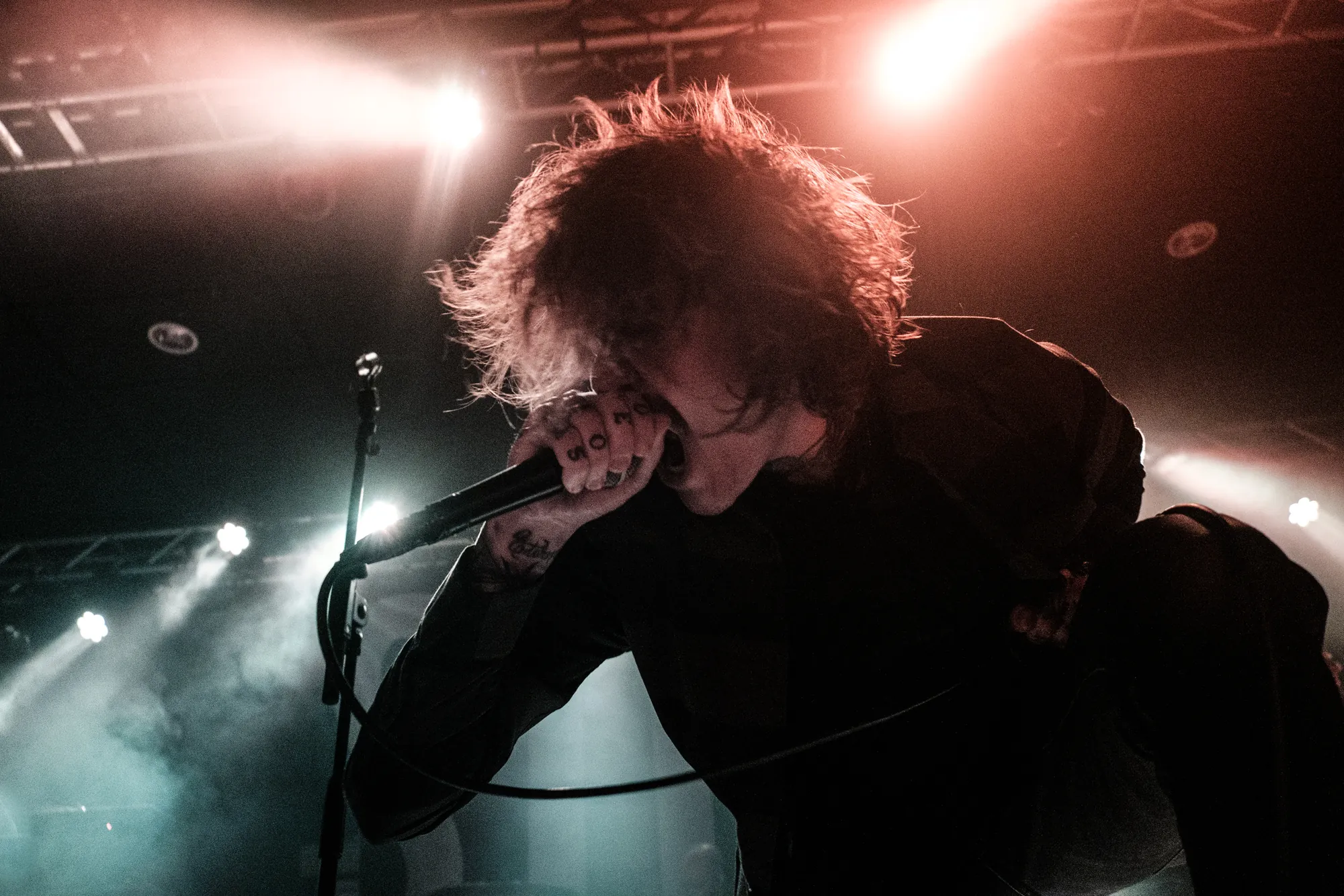 Rock vocalist hunched over the microphone on a dark stage with red and teal light