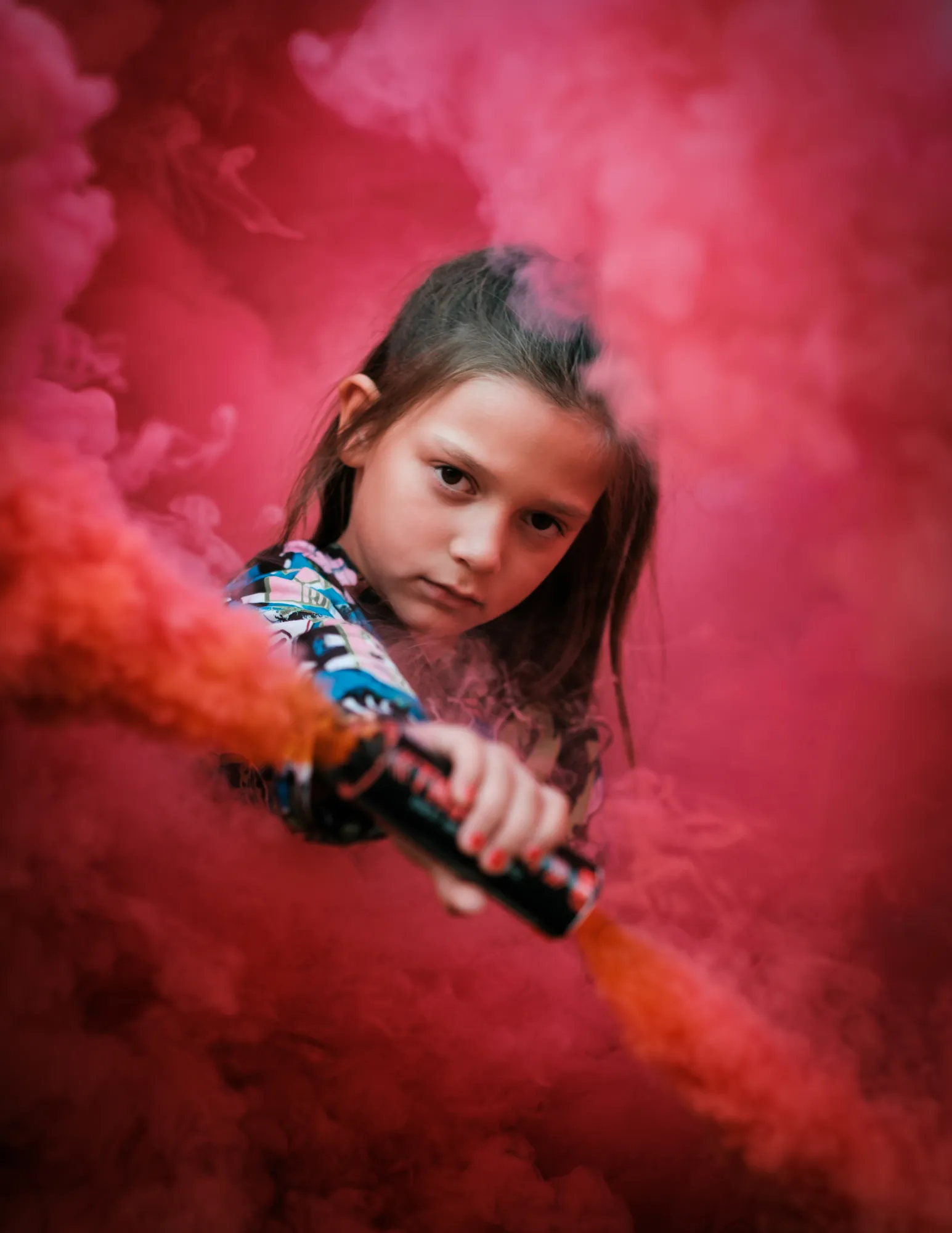 Young girl holding a smoke canister surrounded by vivid red smoke, staring directly at the camera