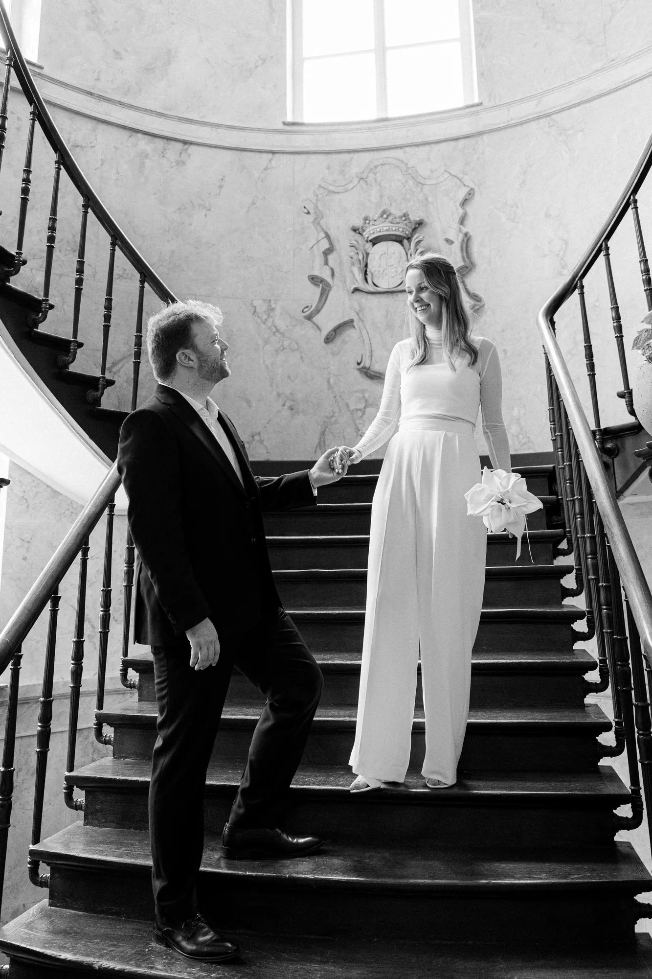 Bride and groom smiling at each other on a grand curved staircase, in black and white