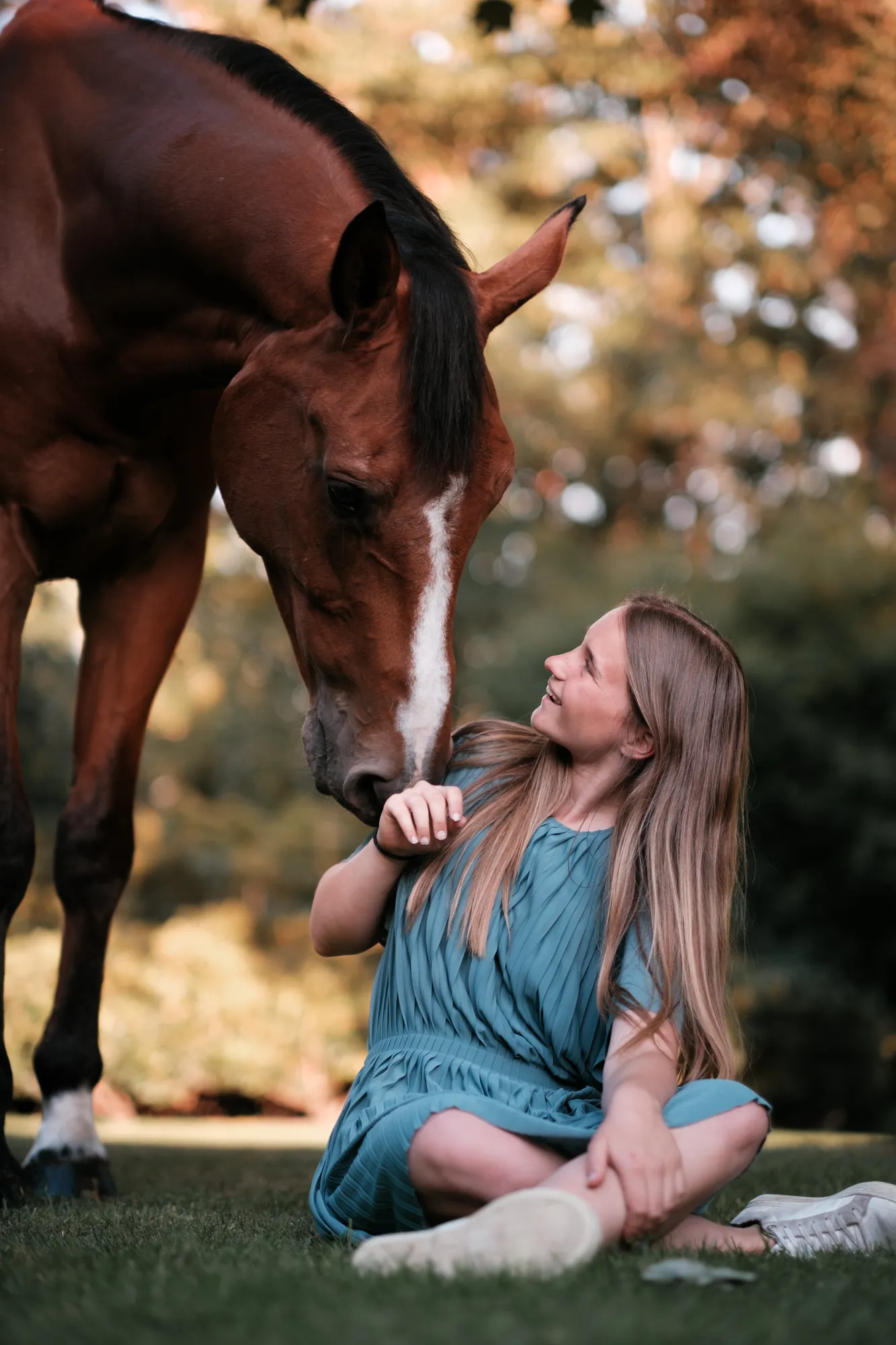 Young woman sitting on grass smiling up at a chestnut horse nuzzling close to her face