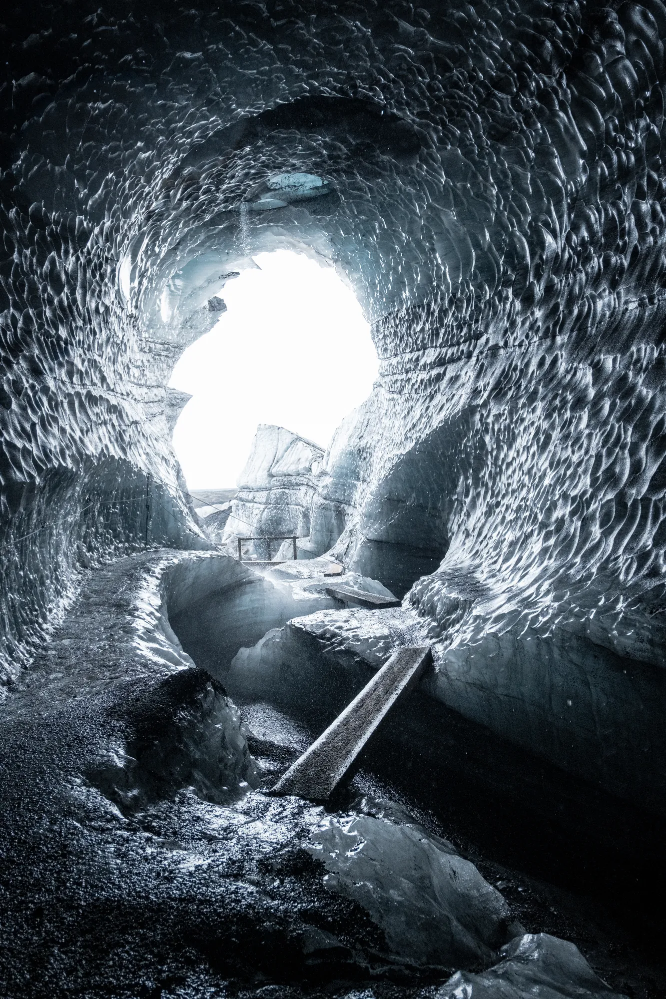 Looking through a glacier ice cave tunnel toward a bright opening, with a stream running through
