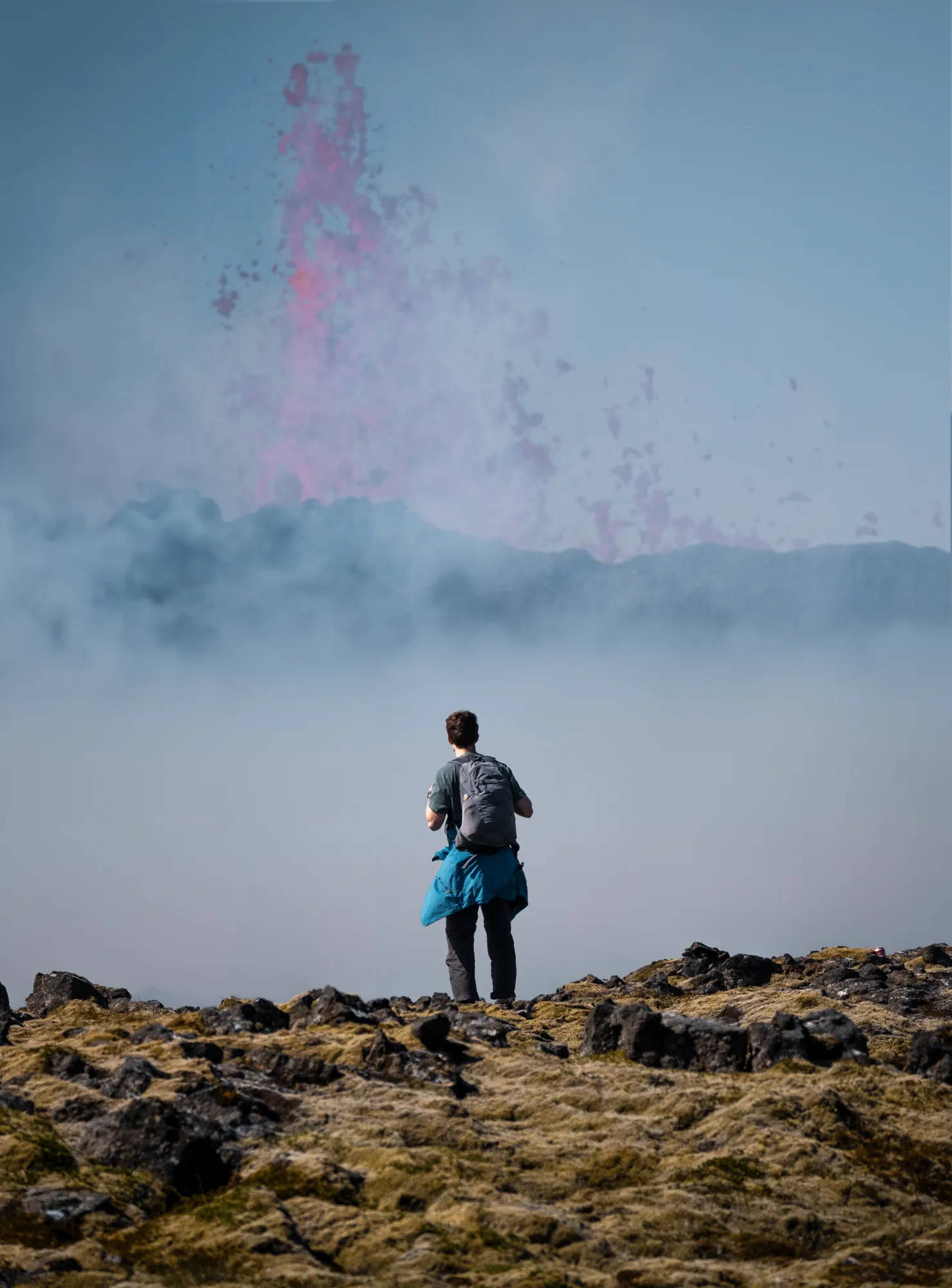 A person standing on a lava field watching a pink eruption plume rise through thick smoke